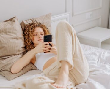 woman in white brassiere lying on bed while holding black mobile phone